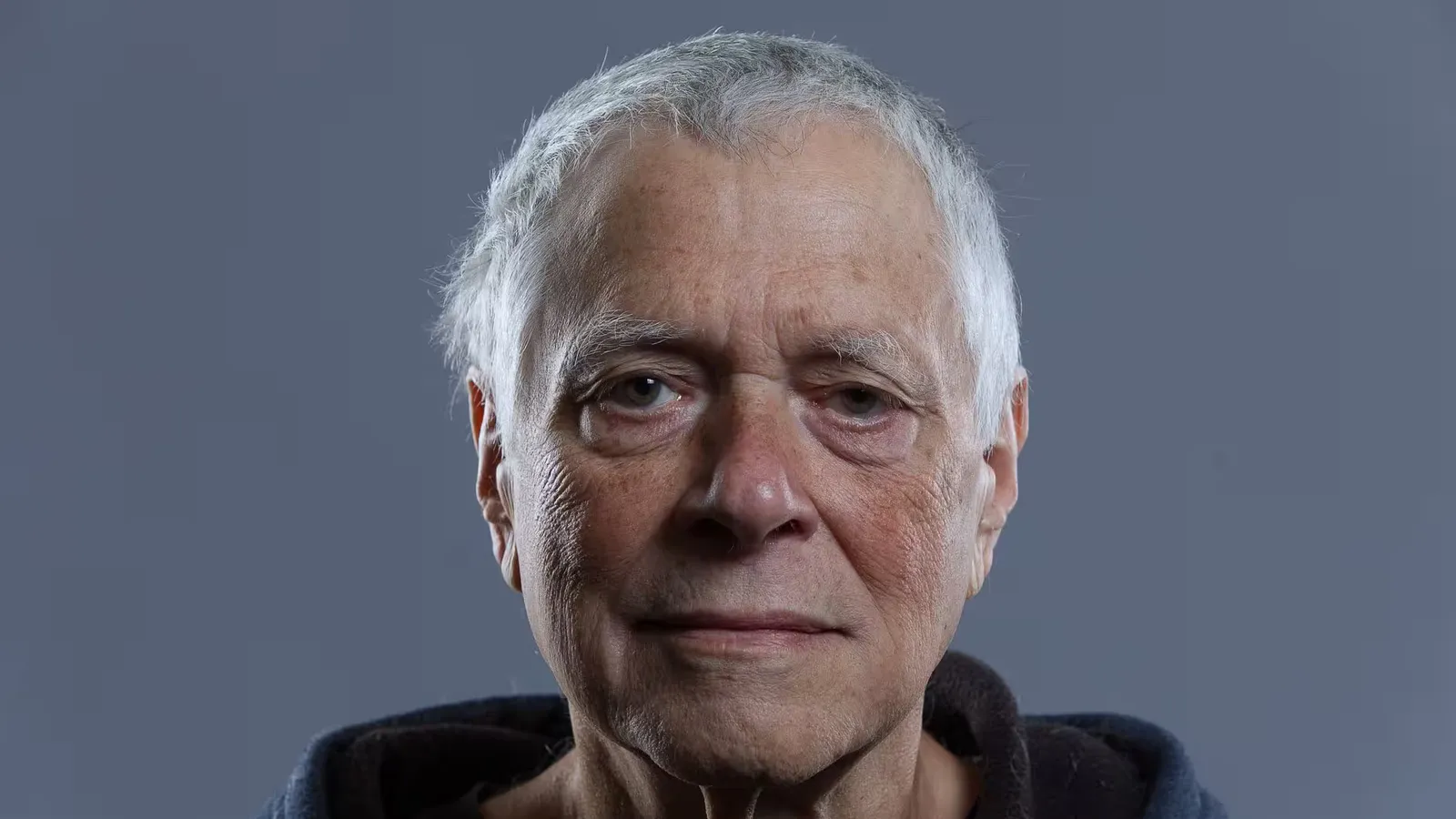 Close-up portrait of an elderly man with a neutral expression, grey background.