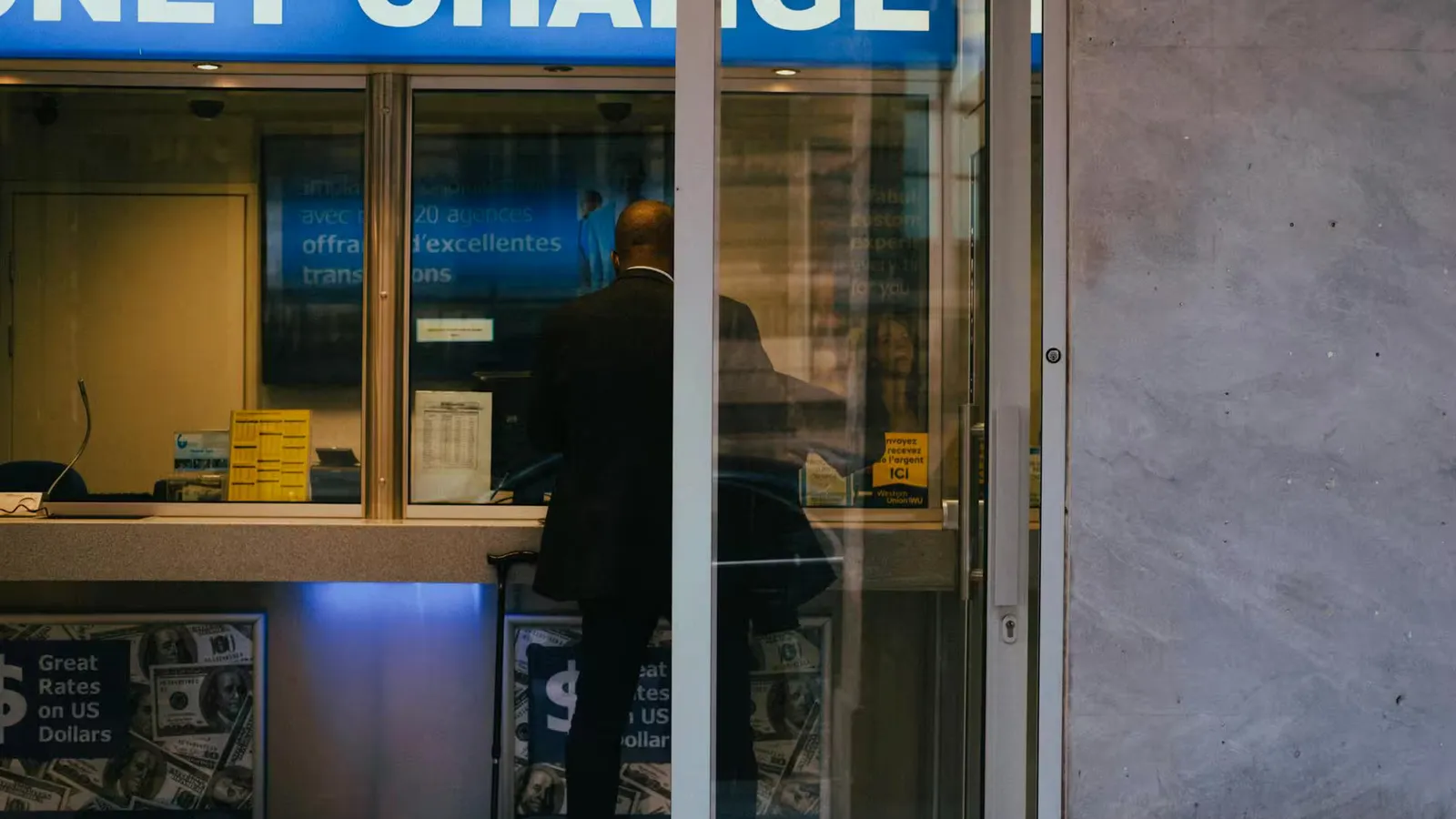 Man at a currency exchange office window, showing currency rates inside a bustling city.