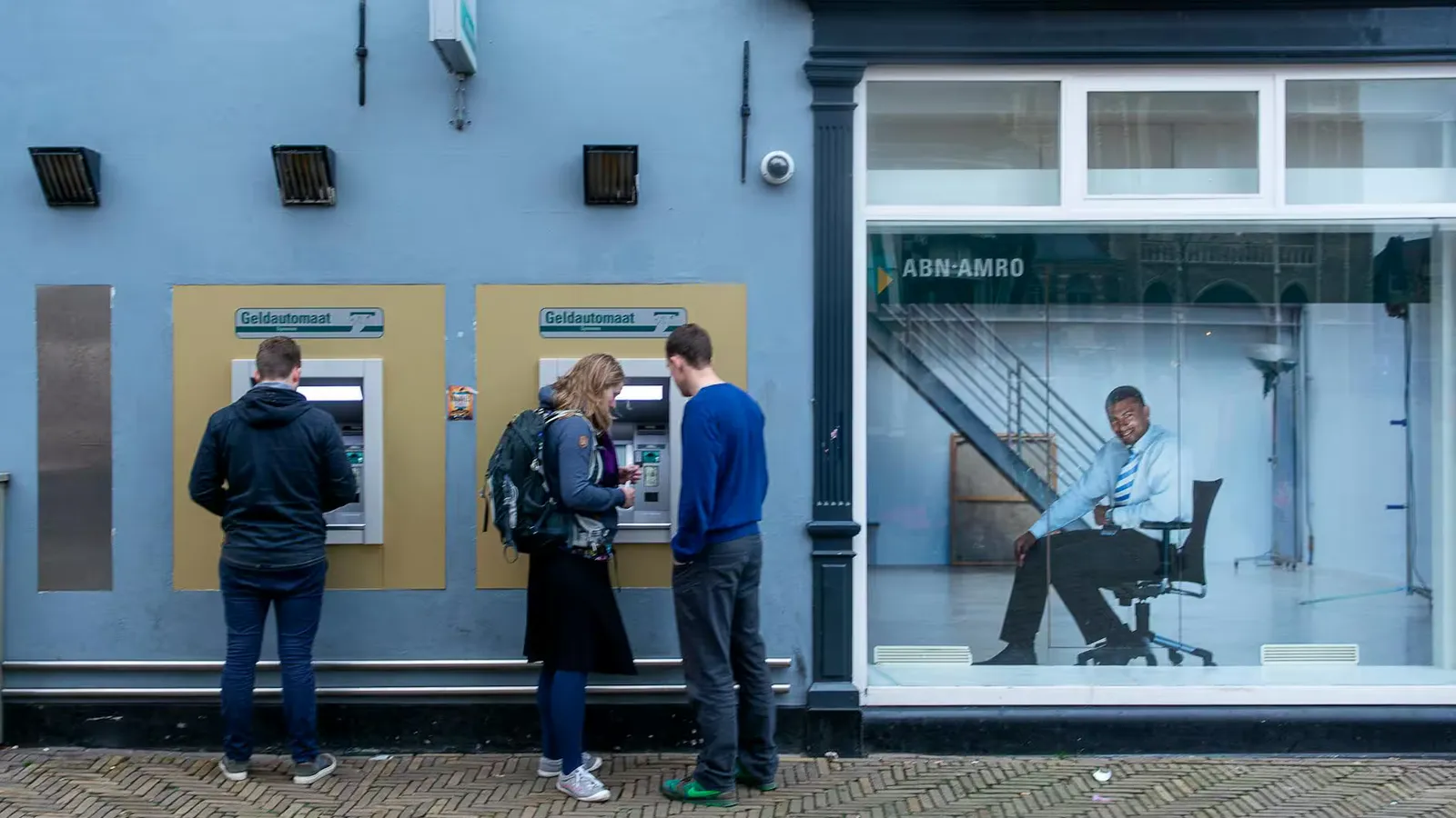 Two ATMs on city street with people using them and bank poster nearby.
