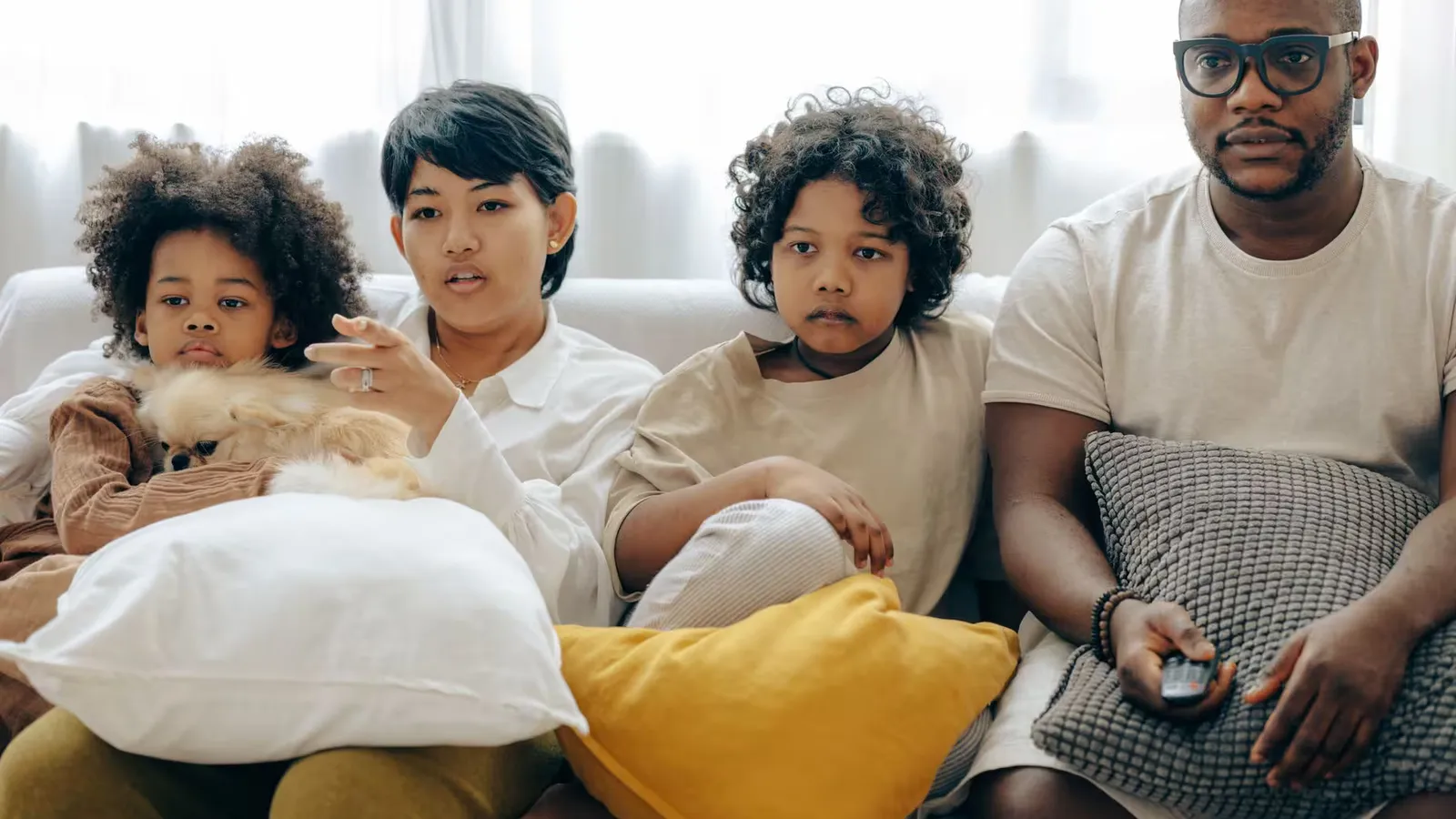 Concentrated multiethnic family watching interesting movie on TV sitting together on cozy couch with different colors pillows and dog in living room while mother pointing at screen