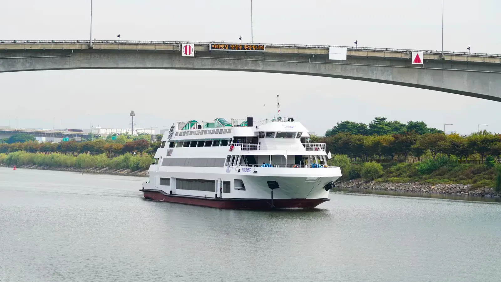 A cruise ship navigates a serene waterway beneath a concrete bridge, set against a lush green landscape.