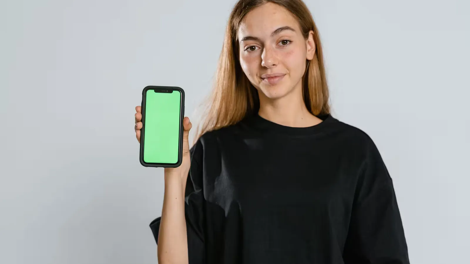 Young woman in black t-shirt showing a green screen smartphone in studio setting, ideal for mockup use.