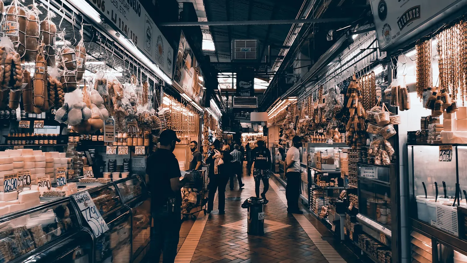 A lively indoor market scene featuring various food stalls and shoppers engaging with vendors.