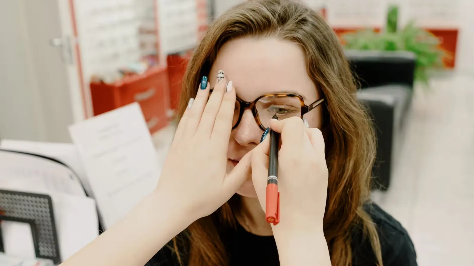 Anonymous female doctor covering eye of female client while choosing proper lenses during appointment in optical salon on blurred background