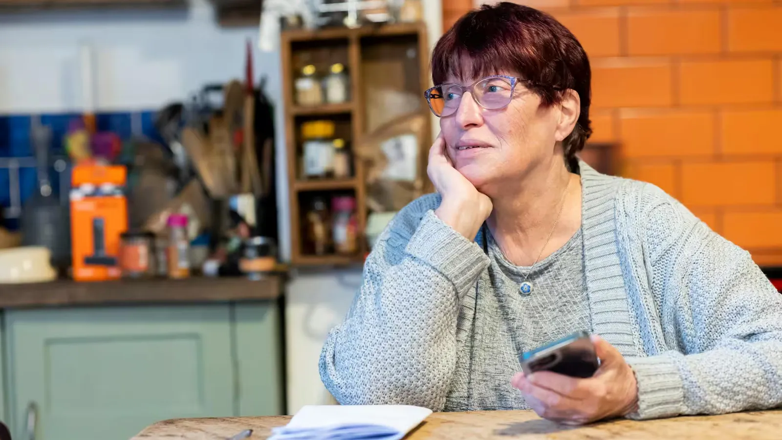 Senior woman in a cardigan pondering while holding a smartphone at a kitchen table.