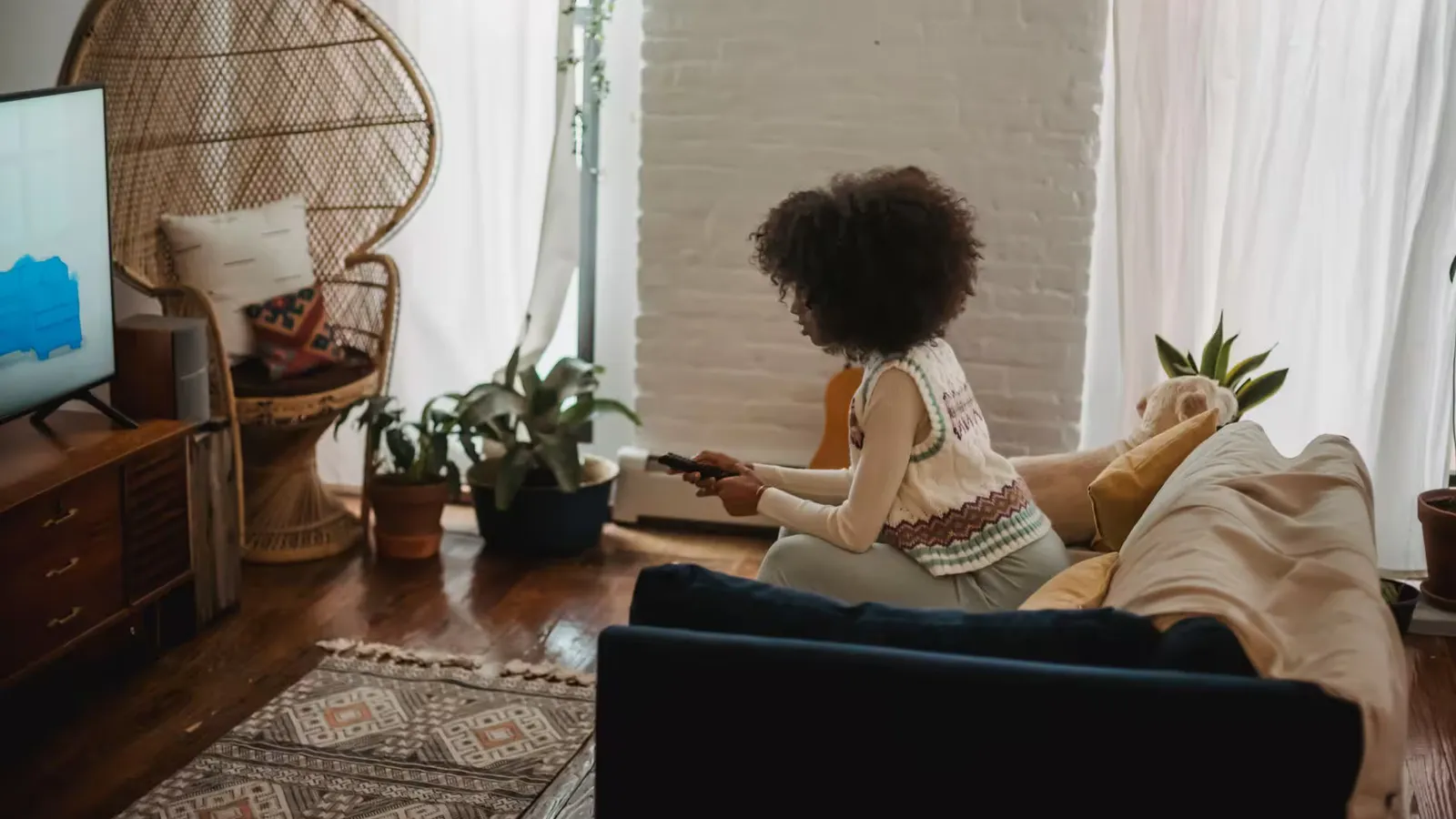 Side view of young African American female millennial with dark curly hair in stylish clothes switching channels on TV with remote controller while resting on sofa during weekend at home