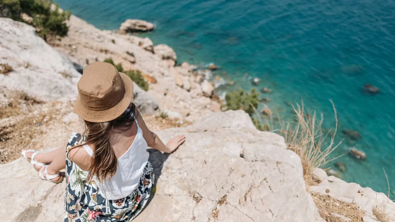 A woman sits on a rocky cliff, enjoying the summer view of turquoise waters.