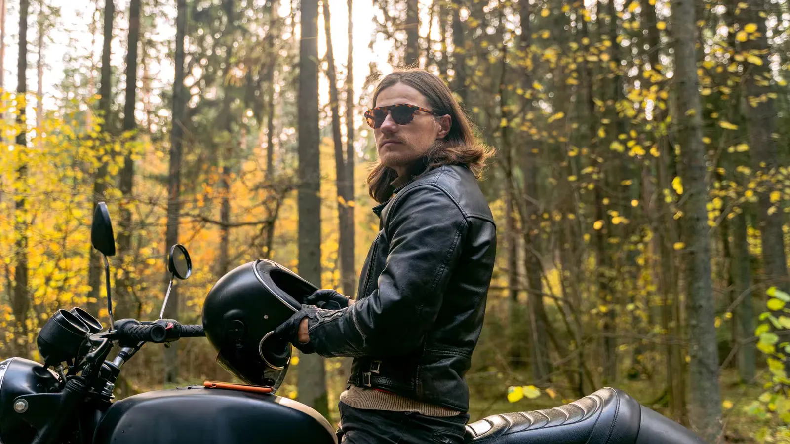 Man in black leather jacket with a motorcycle helmet in an autumn forest setting.
